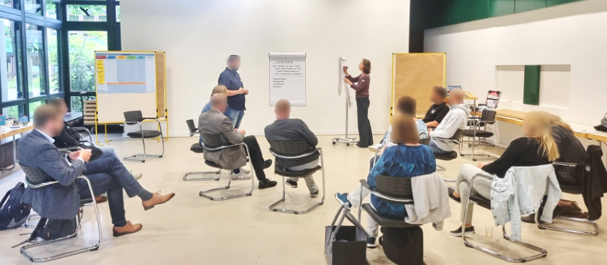 Workshop-Situation mit Vortragenden, Person am Flipchart, Stuhlkreis